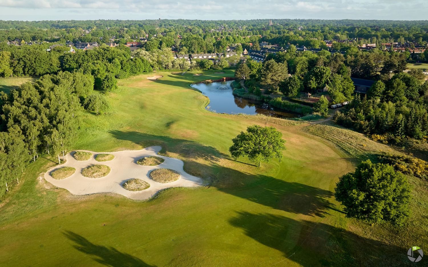 Aerial view of a lush golf course featuring sand bunkers, a pond, and tree-lined fairways on a sunny day.