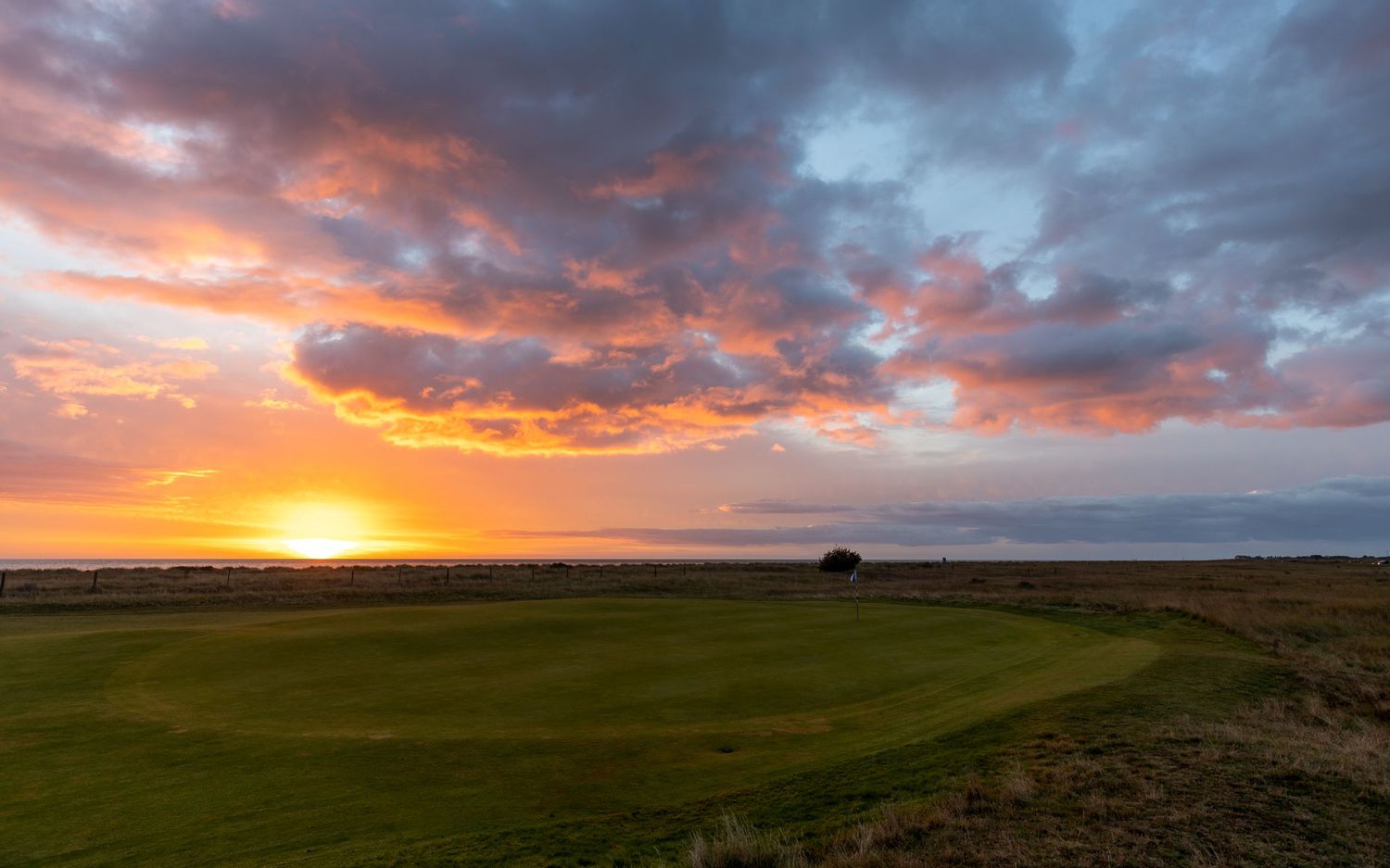 Stunning sunset over a golf course green with dramatic orange and blue clouds reflecting over the ocean horizon.