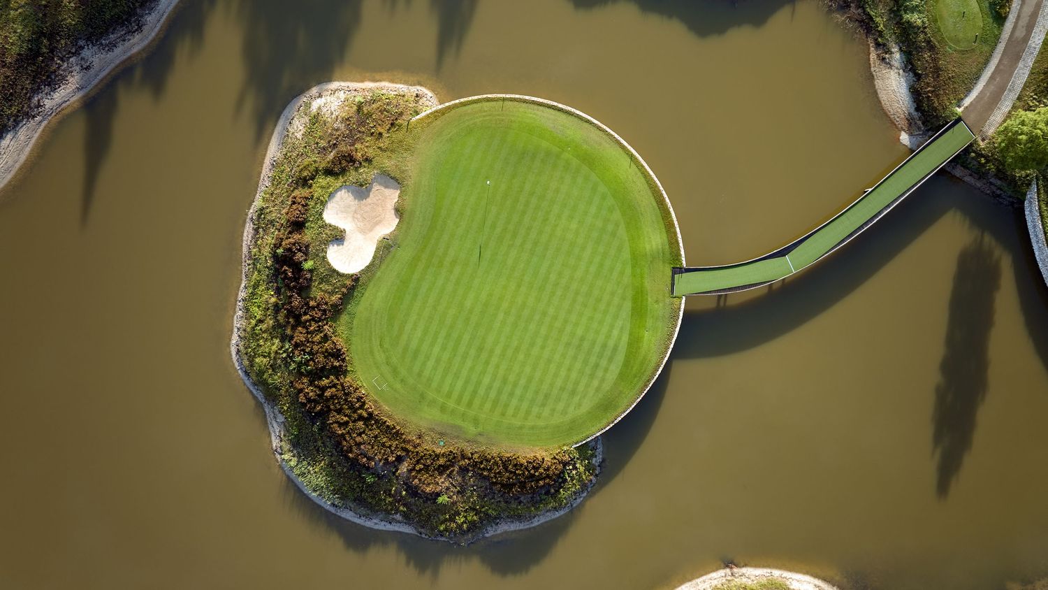 Aerial view of an island golf green surrounded by water hazard with a narrow bridge pathway.