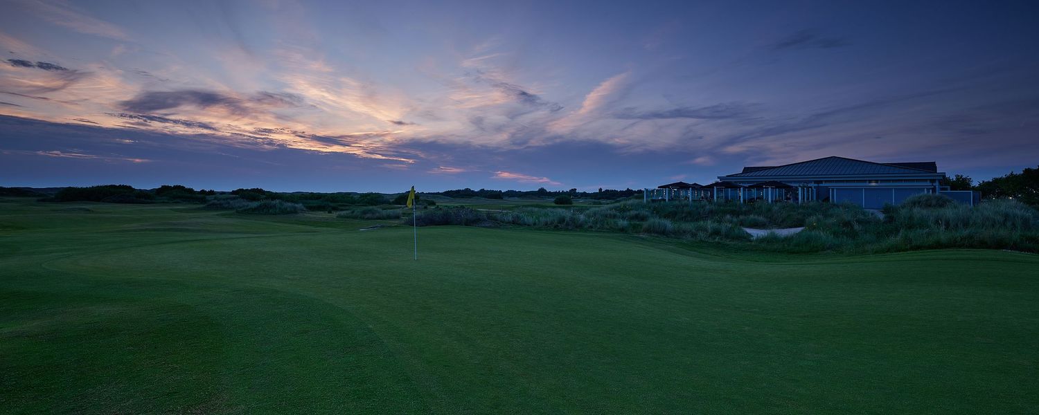 Golf course at dusk with dramatic sunset sky, green fairway, and clubhouse silhouette in background.