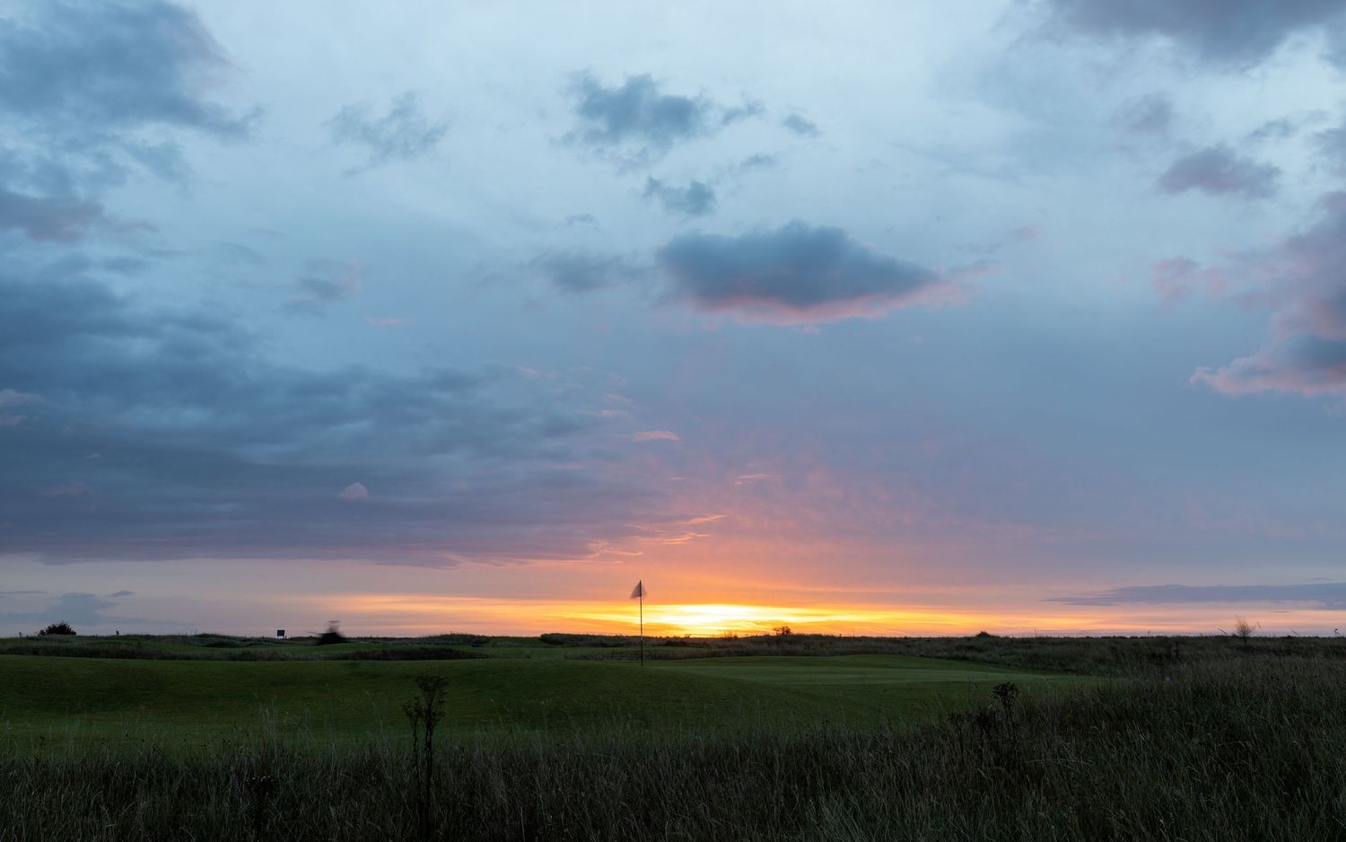 Serene golf course sunset landscape with a distant flagstick silhouetted against a vibrant golden horizon under vast skies.