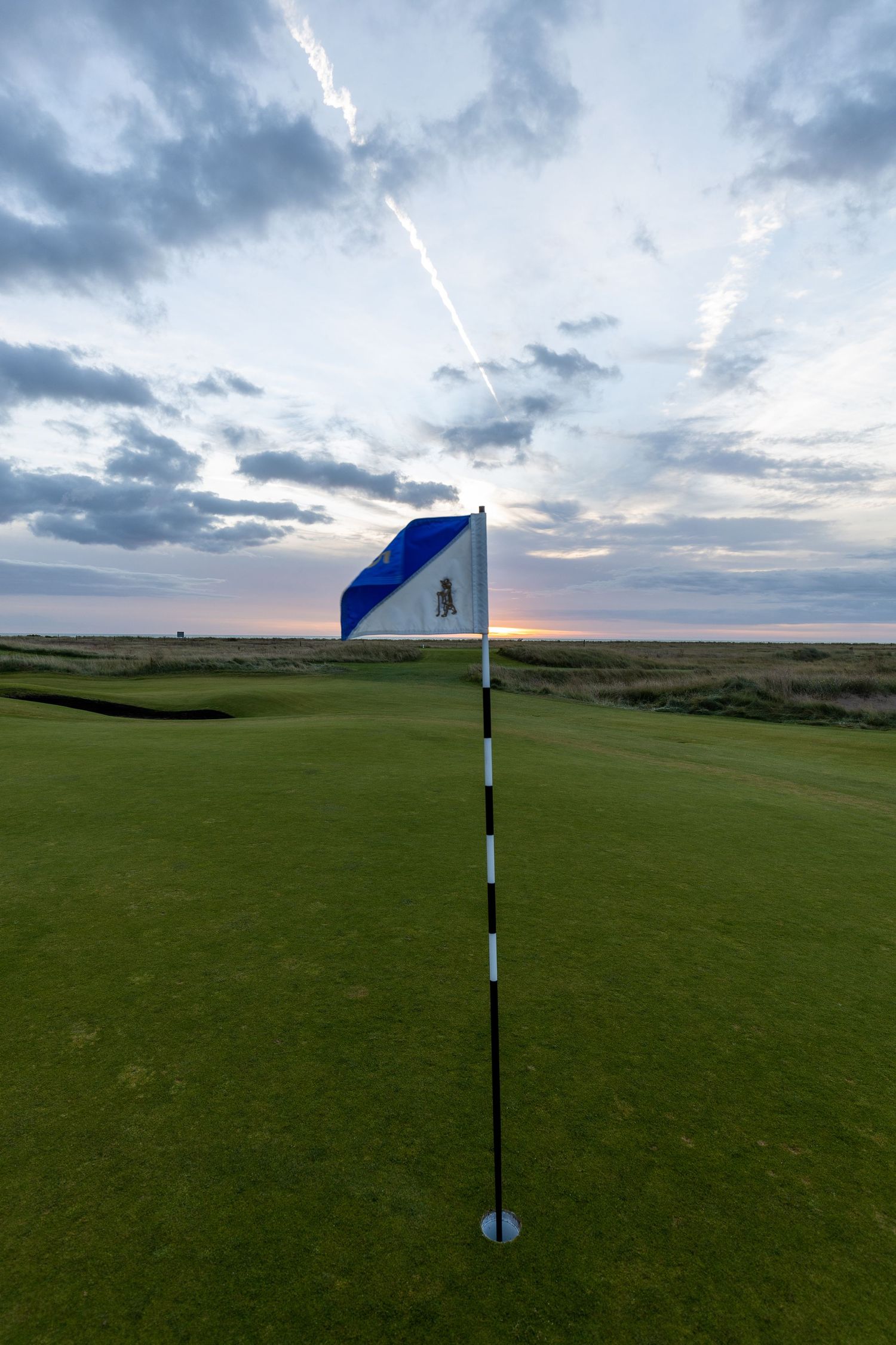 Golf course green at sunset with a blue and white flag on a black and white striped pin under a dramatic cloudy sky.
