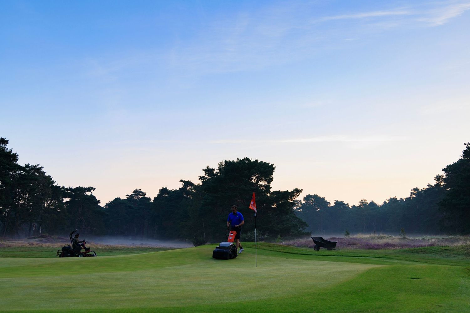 Early morning golfers prepare to tee off on a misty golf course with golf carts visible in the distance.