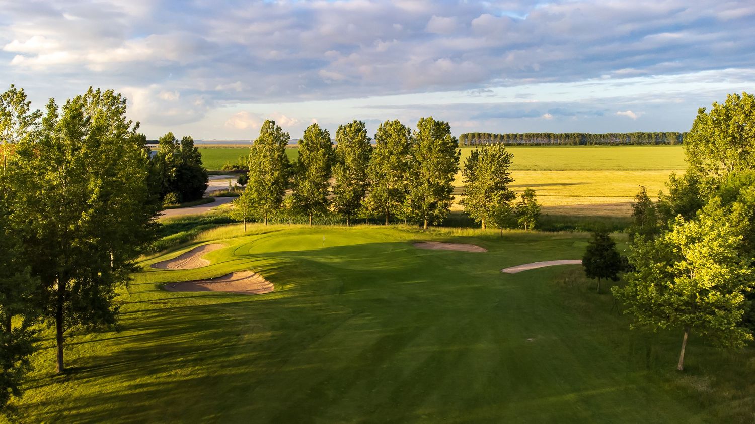 A panoramic view of a lush golf course with rolling fairways, scattered trees and dramatic clouds in the sky.