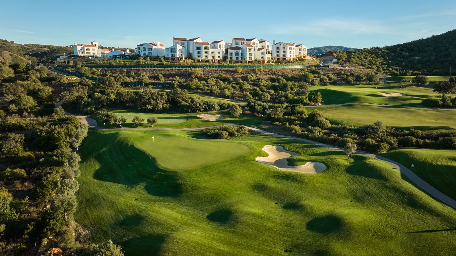 Panoramic view of a lush green golf course with rolling hills, sand bunkers, and luxury residences in the background.