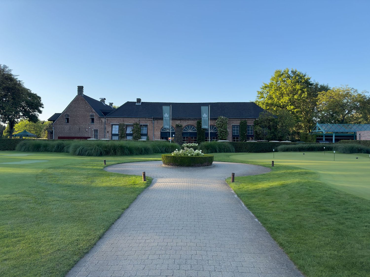 Brick clubhouse with paved pathway, circular garden, and lush green lawn under clear blue sky.