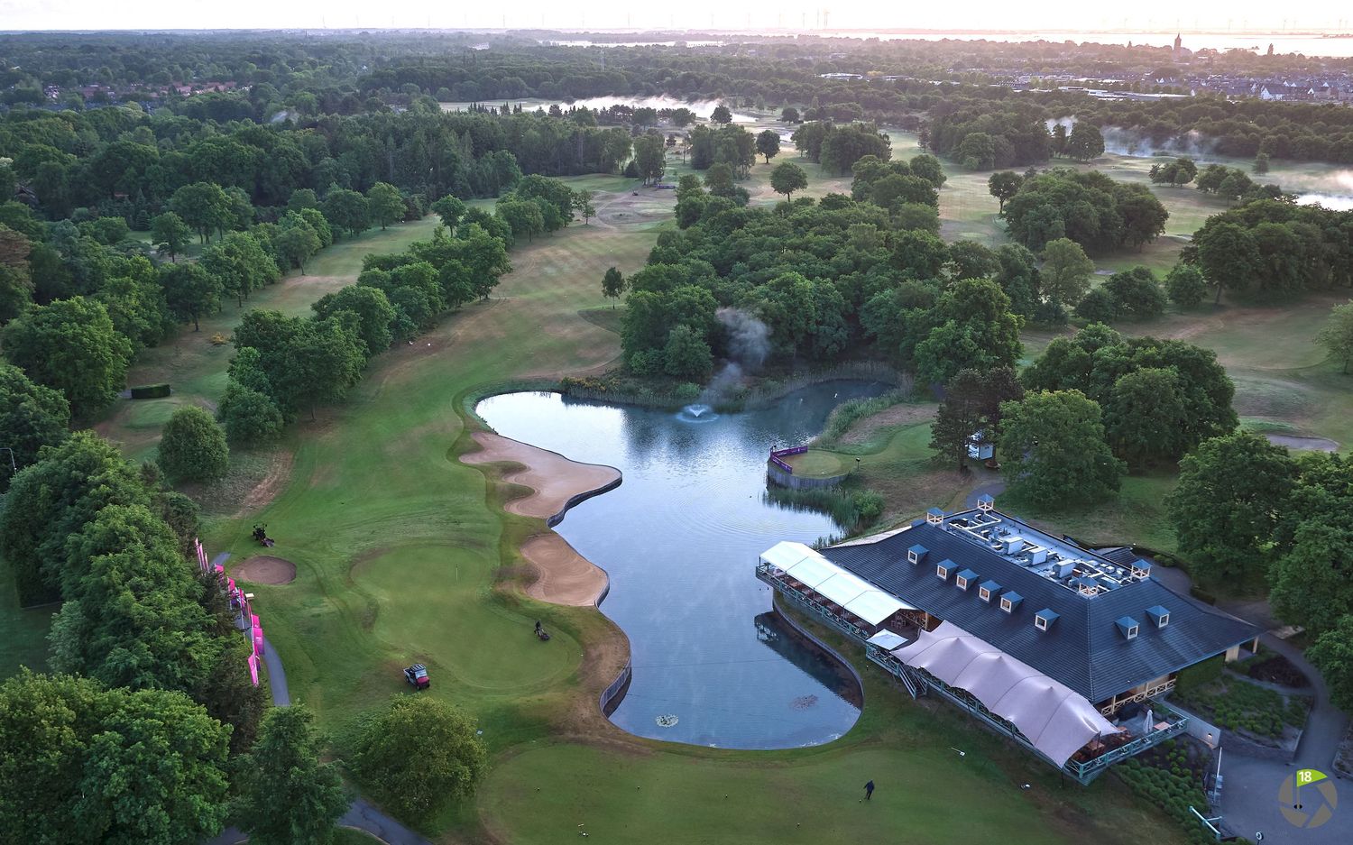 Aerial view of a lush golf course with a scenic pond, clubhouse, and city skyline at dusk.
