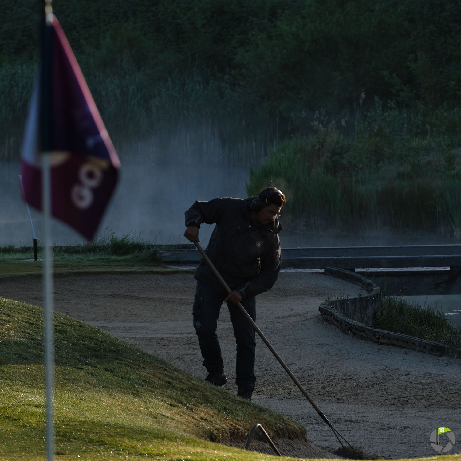Groundskeeper raking a sand bunker on a misty golf course at dusk near a flagged hole.