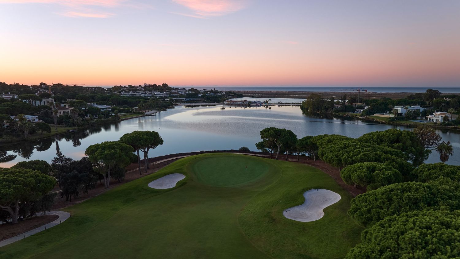 Twilight aerial view of a golf course with sand bunkers, a calm lake, and a colorful pink and purple sunset sky.