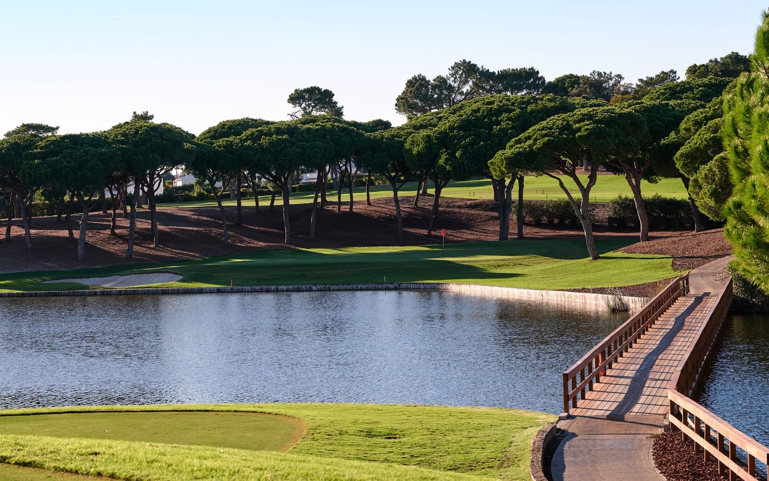 Scenic golf course with a tranquil lake, wooden bridge, and tall pine trees under a clear blue sky.