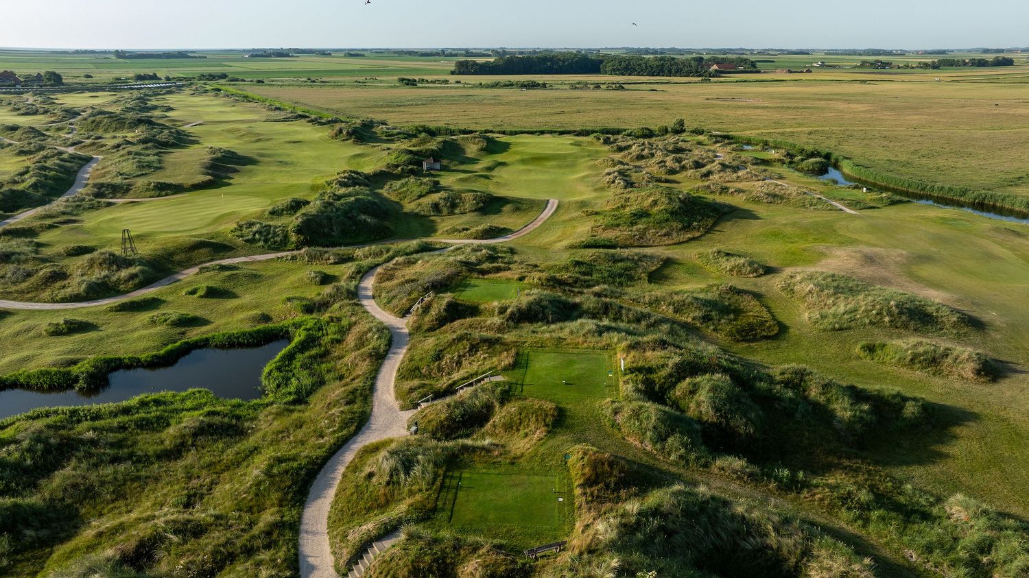 Aerial view of a links golf course with winding paths, green fairways, and natural dune landscape under open skies.