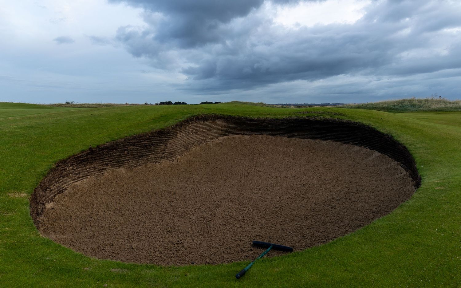 Large deep sand bunker on a golf course fairway under dramatic cloudy skies.