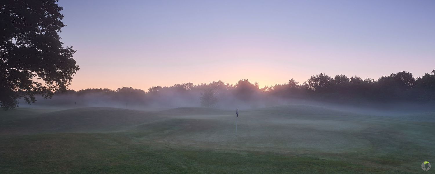 Misty golf course at sunrise with a lone flag on the green, surrounded by trees in soft purple and pink morning light.