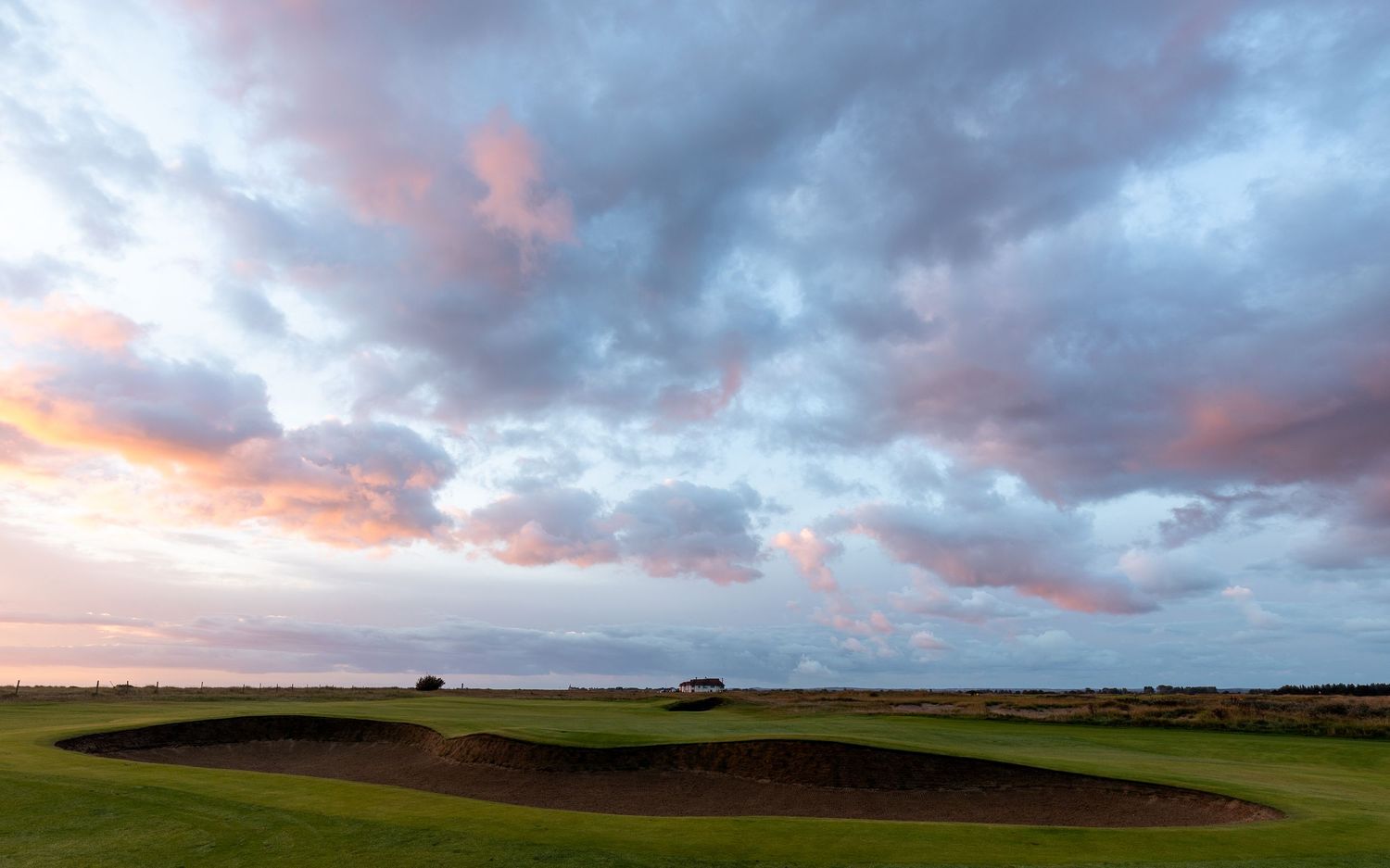 Dramatic pink and blue sunset clouds over a links golf course with a prominent sand bunker.