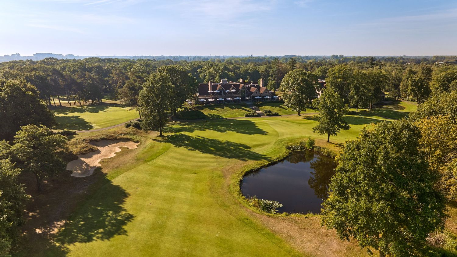 Aerial view of a golf course with a pond, bunkers, and a grand clubhouse amid trees.