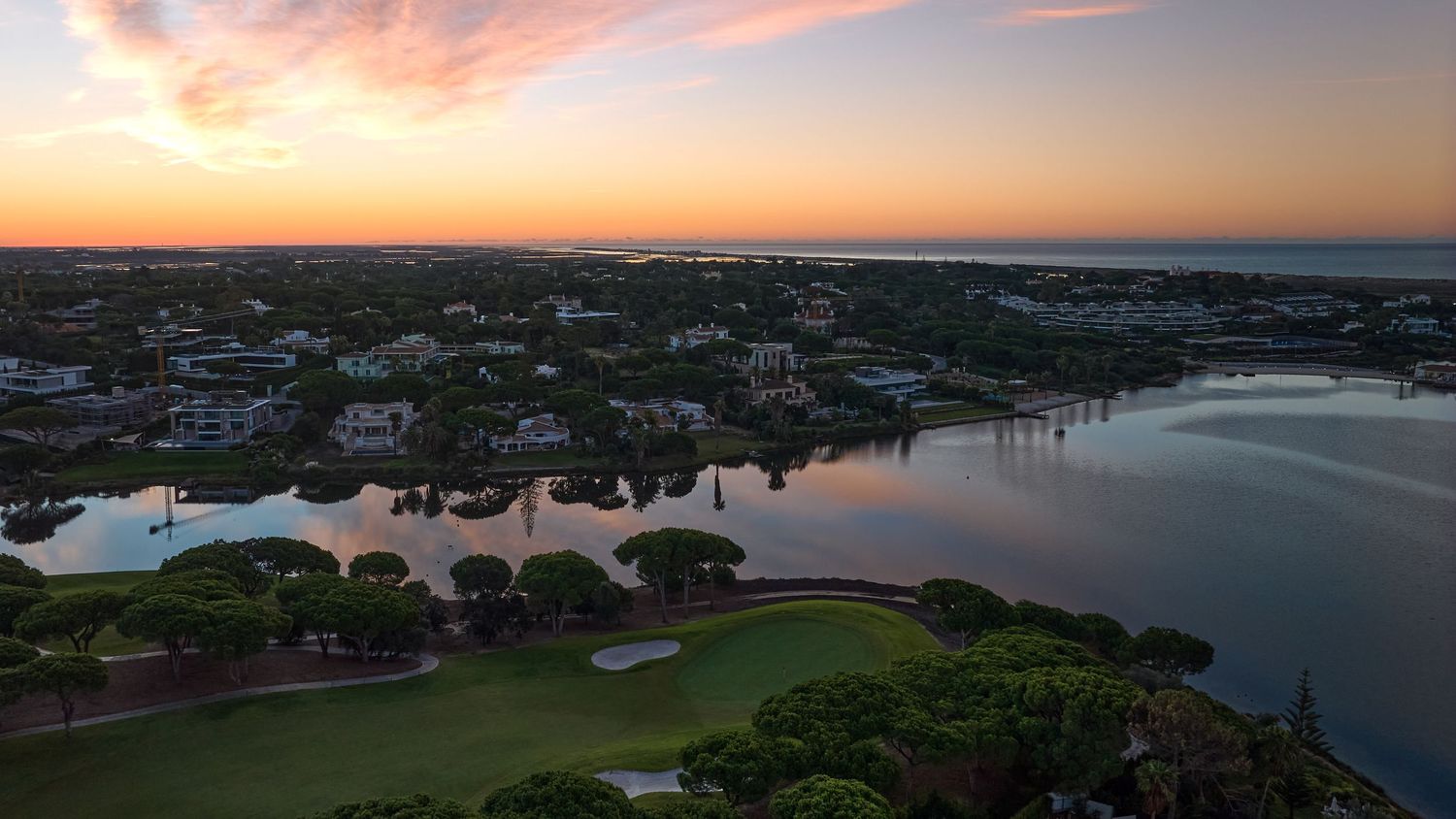 Aerial sunset view of a coastal golf course community with calm water reflections and vibrant orange skies.