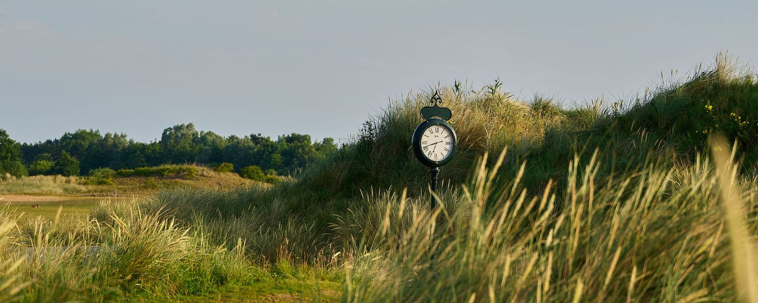 A classic clock stands amid tall grass dunes on a scenic golf course under a clear sky.