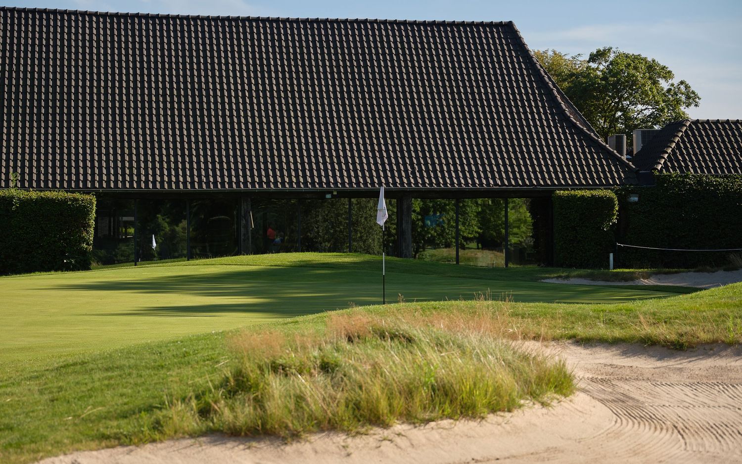 Golf course green with sand bunker near a traditional clubhouse building with dark tiled roof.