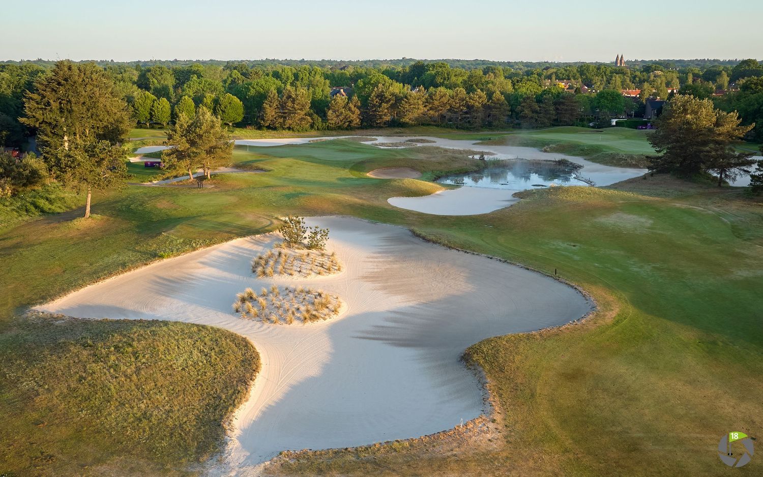 Aerial view of a scenic golf course featuring large sand bunkers, water hazards, and lush green fairways surrounded by trees.