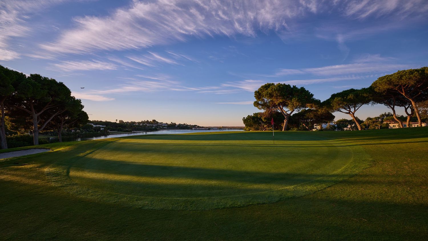 Lush green golf course fairway at sunset with tall pine trees and a serene blue sky in the background.