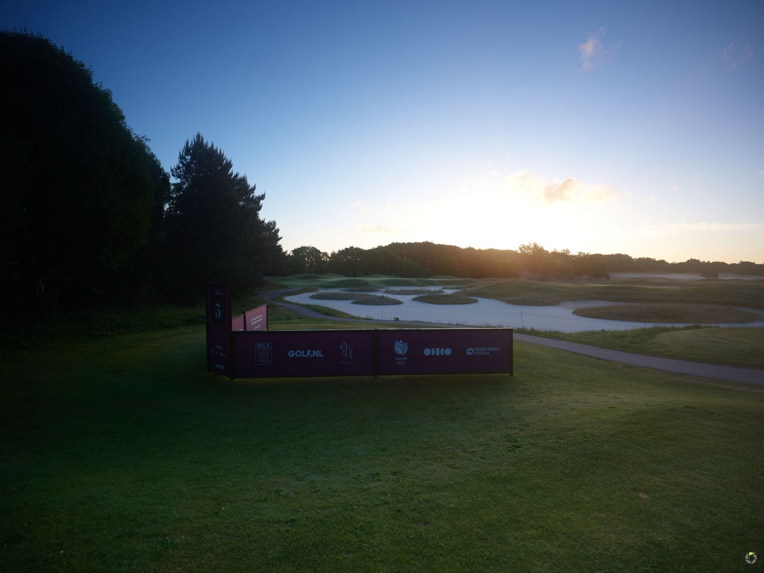 Sunset behind a golf course scoreboard with a serene water hazard and tree-lined fairway in the background.