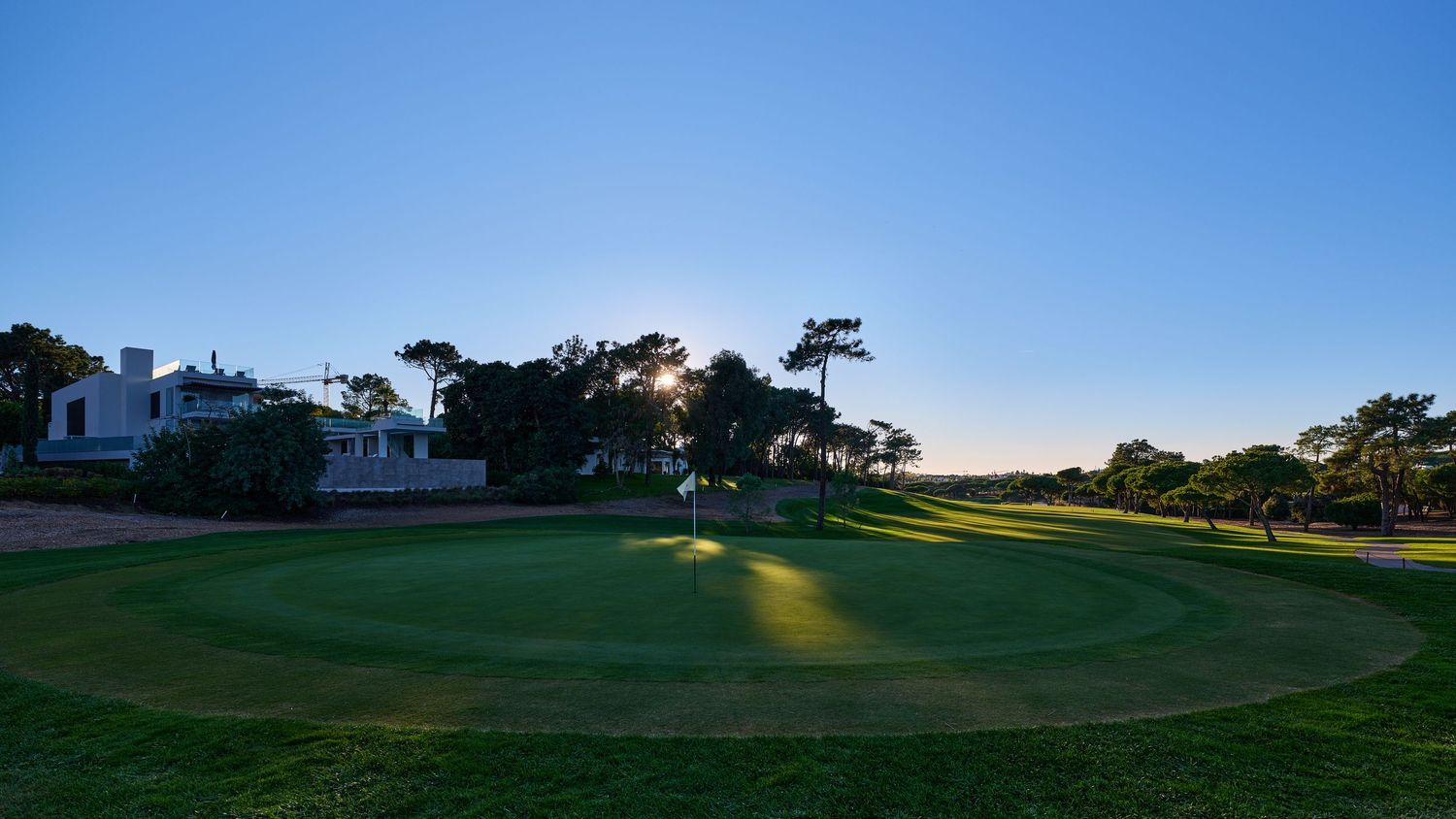 Scenic golf course green at sunset with lush fairways, trees, and a clear blue sky in the background.