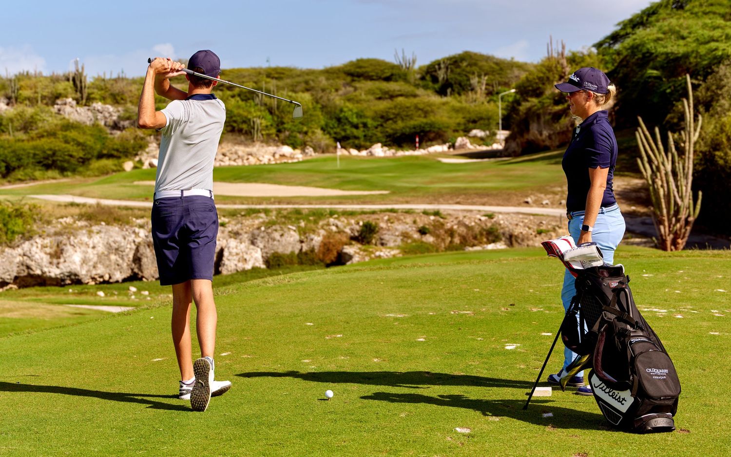 Golfers practicing their swings on a sunny course with golf bags and desert landscape.