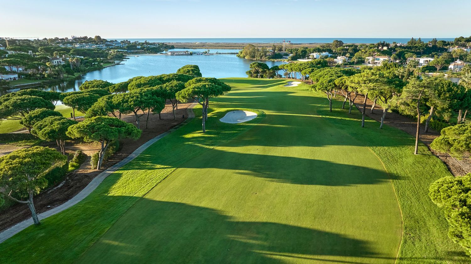 Aerial view of a lush green golf course with sand bunkers, pine trees, and a scenic lake in the background.