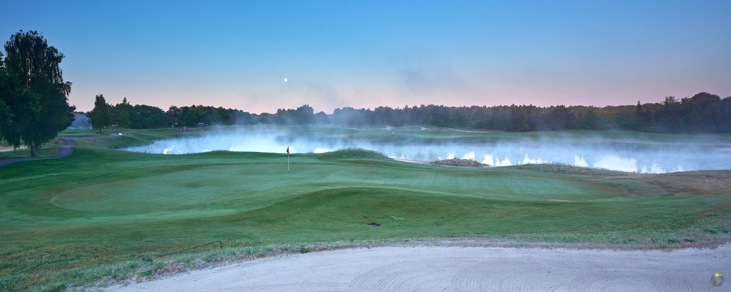 Misty early morning view of a lush green golf course fairway with fog rolling over the grass under a soft blue sky.