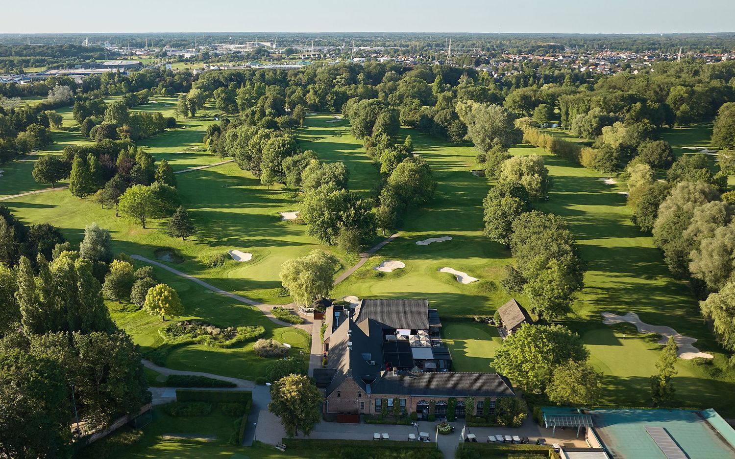 Aerial view of a lush green golf course with sand bunkers, mature trees, and a clubhouse surrounded by a scenic town.