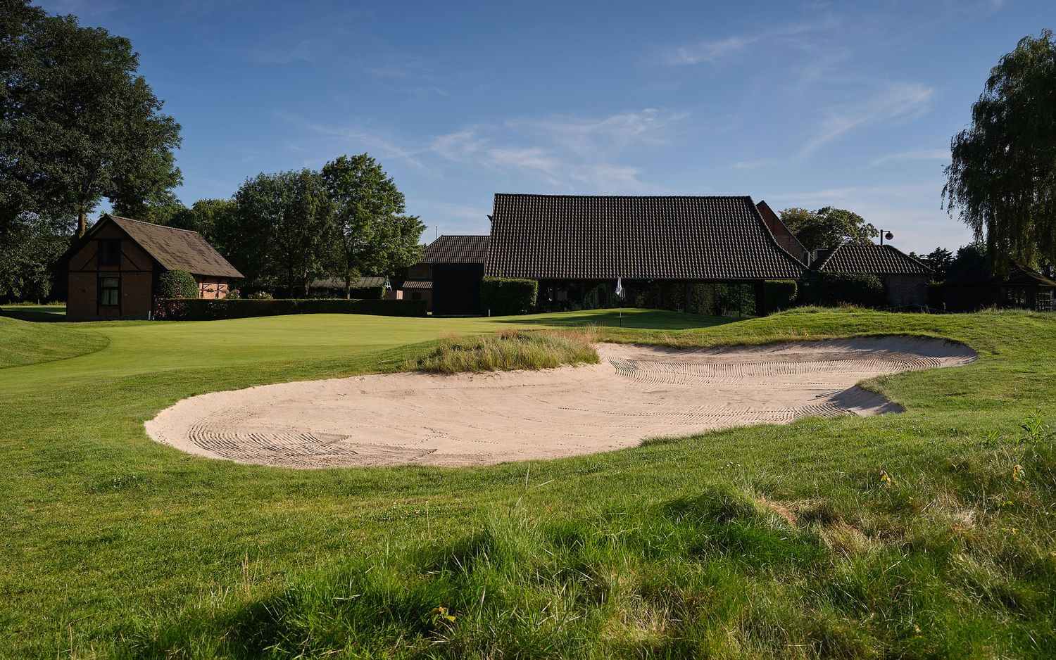 Golf course sand bunker beside a well-maintained fairway with traditional clubhouse buildings in the background.