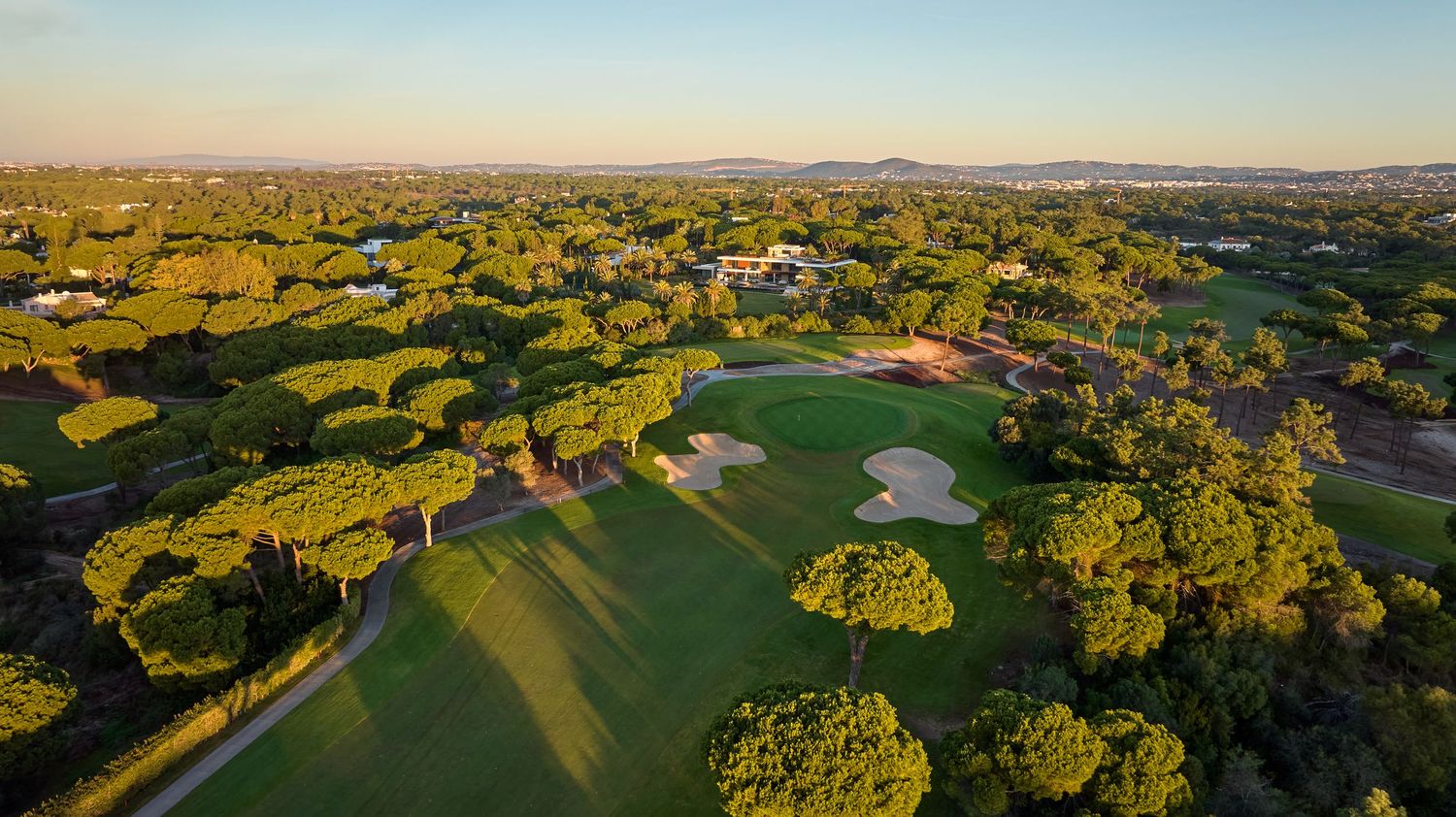 Aerial view of a lush green golf course surrounded by dense trees at golden hour in a scenic landscape.