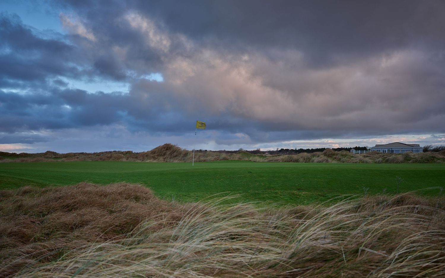 A windswept links golf course with a yellow flag, wild grass dunes, and dramatic stormy clouds at dusk.