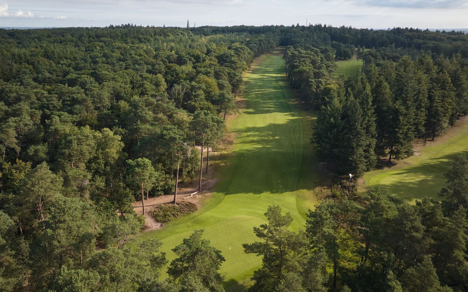 Aerial view of a narrow golf fairway cutting through dense green forest under an overcast sky.
