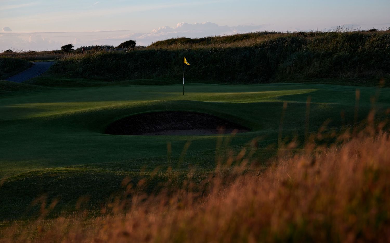 Links golf course at dusk with yellow flag, sand bunker, and golden fescue grass in soft evening light.