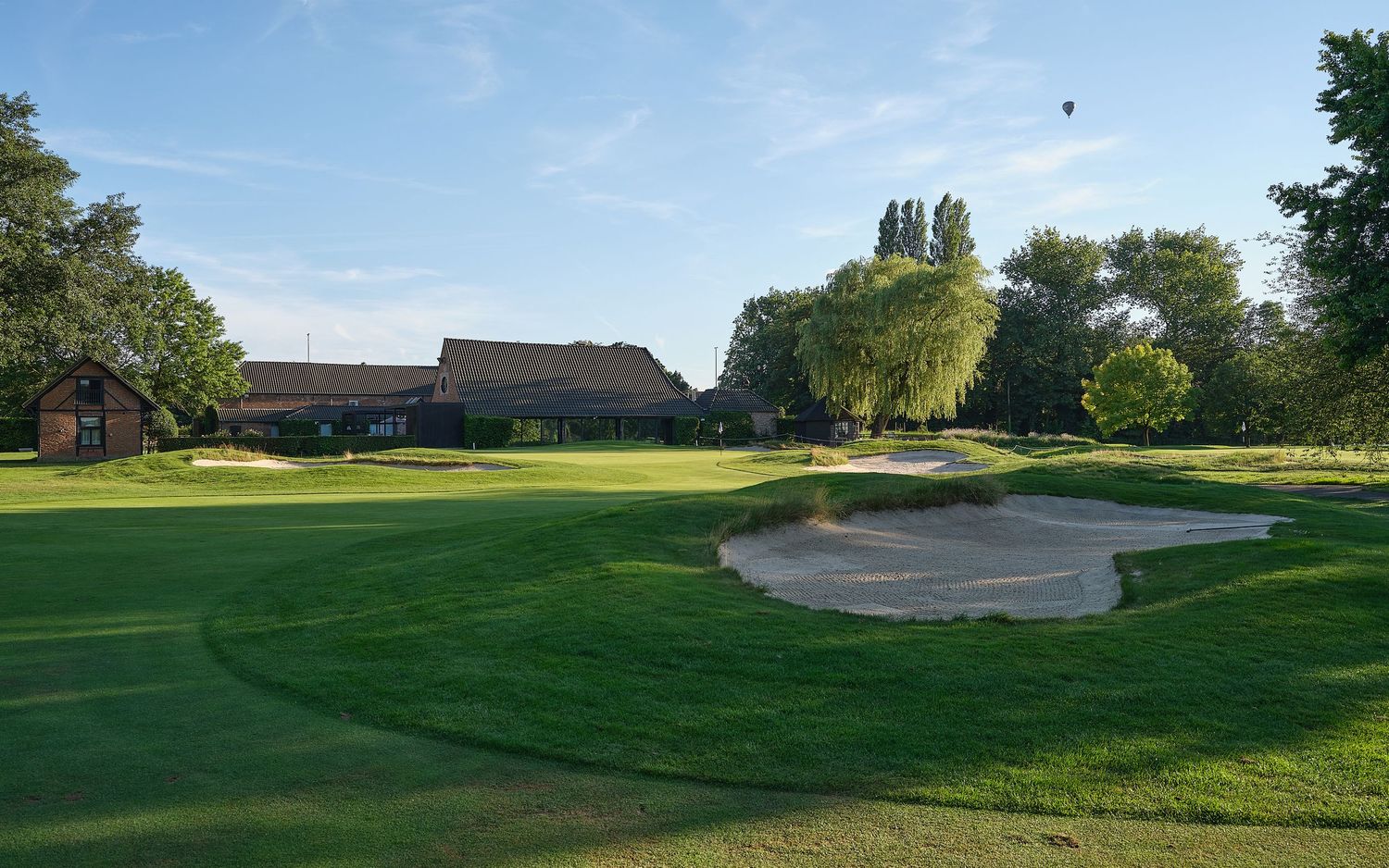 Golf course fairway with large sand bunker near the clubhouse building surrounded by mature trees on a sunny day.