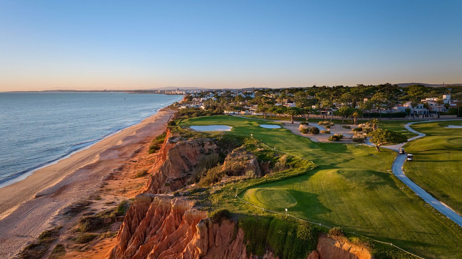 Panoramic aerial view of a coastal golf course with dramatic cliffs overlooking the ocean at sunset.