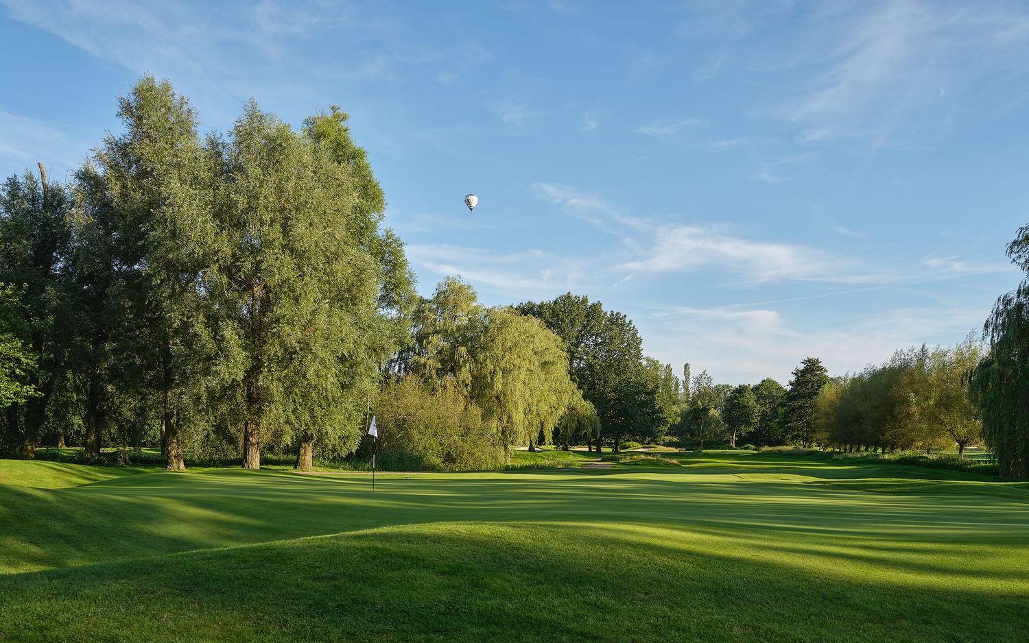 Lush green golf course with willow trees under a blue sky, featuring a hot air balloon floating in the distance.