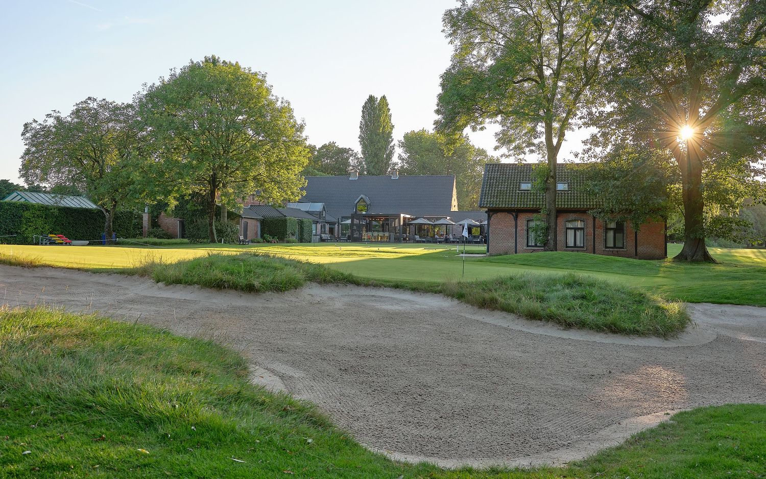 Golf course clubhouse surrounded by mature trees with a sand bunker in the foreground at golden hour.