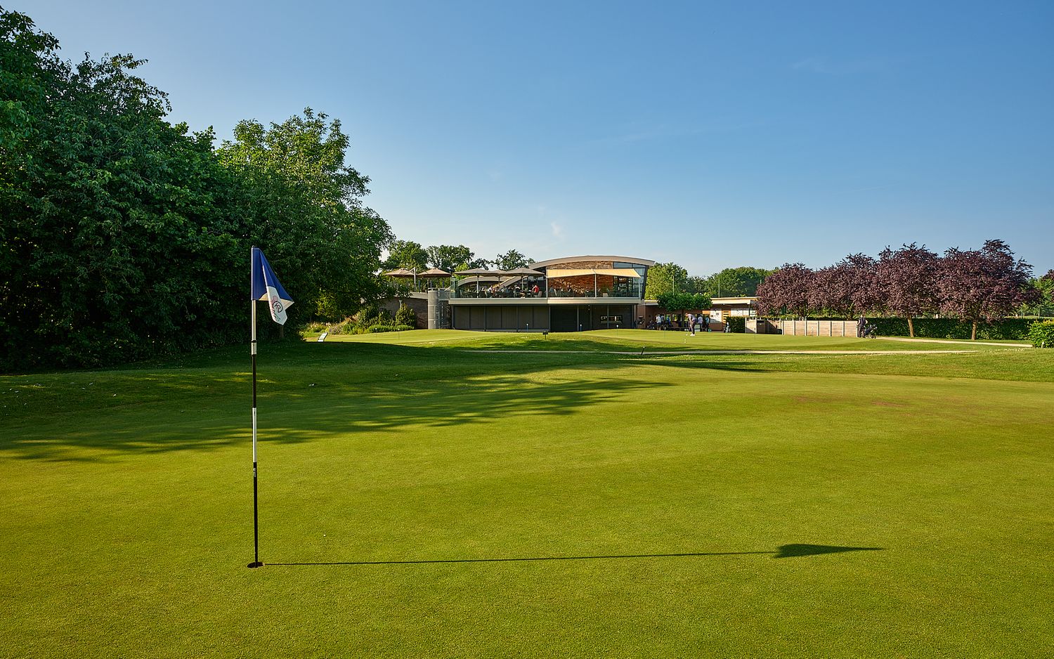 A scenic golf course green with a flag pin in the foreground and a clubhouse building visible on the hill in the background.