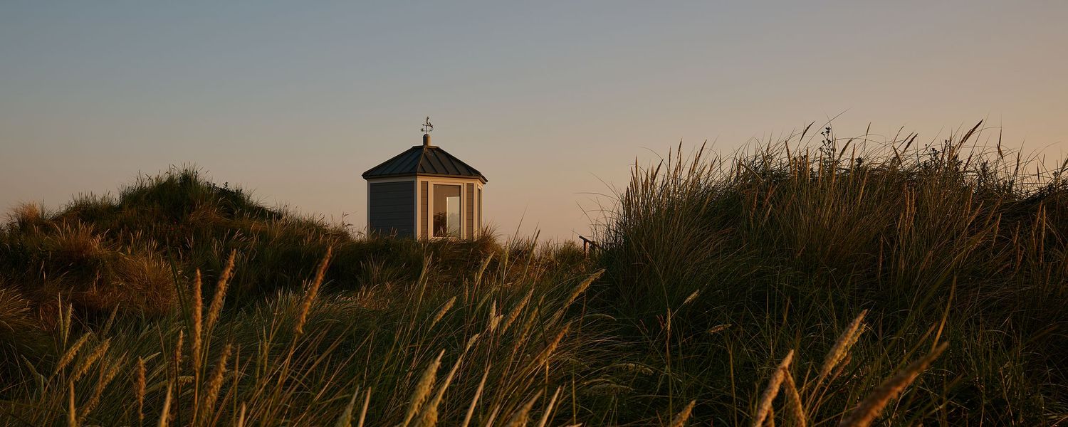 Small white chapel sits atop sandy dunes surrounded by tall golden grass at sunset on a coastal landscape.