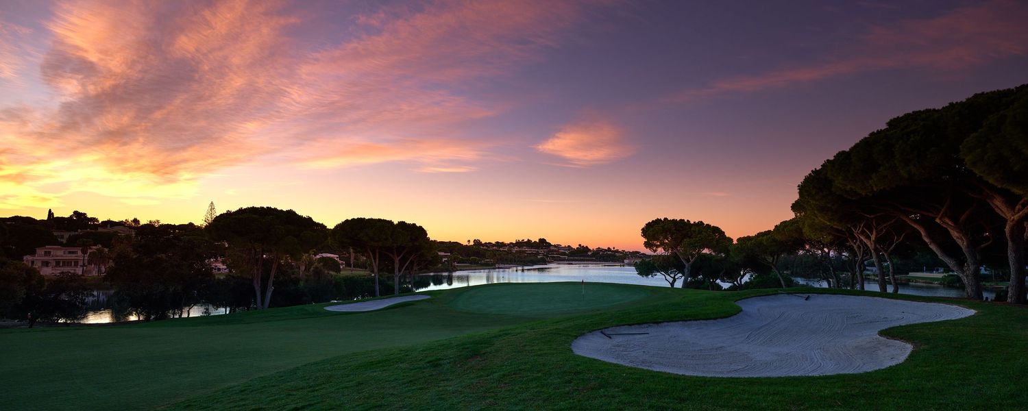 Panoramic view of a golf course with sand bunkers and trees silhouetted against a dramatic purple and orange sunset sky.