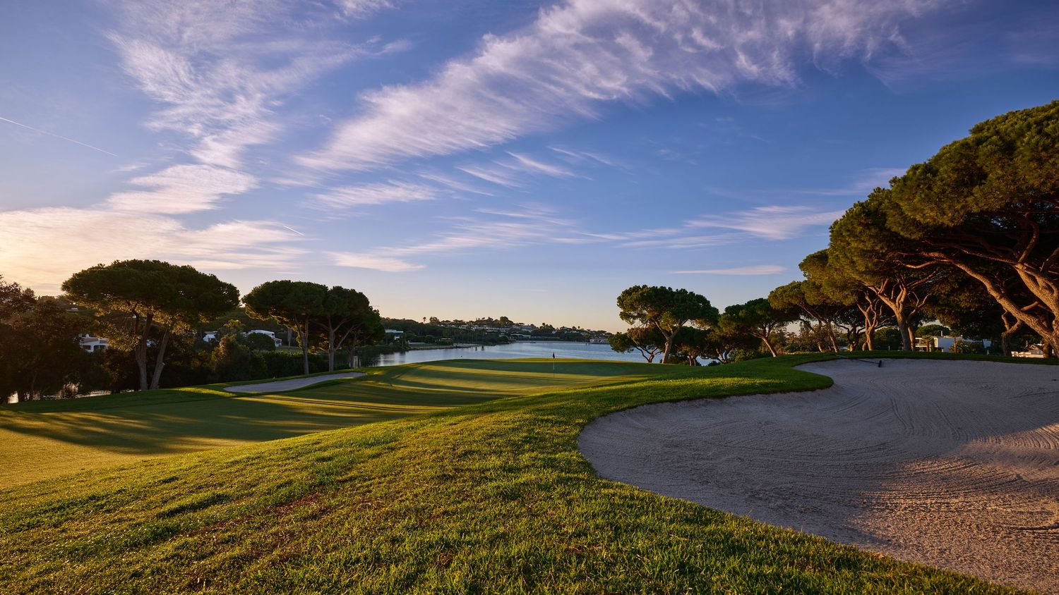 Serene golf course with water hazards and pine trees silhouetted against a pastel sunset sky.