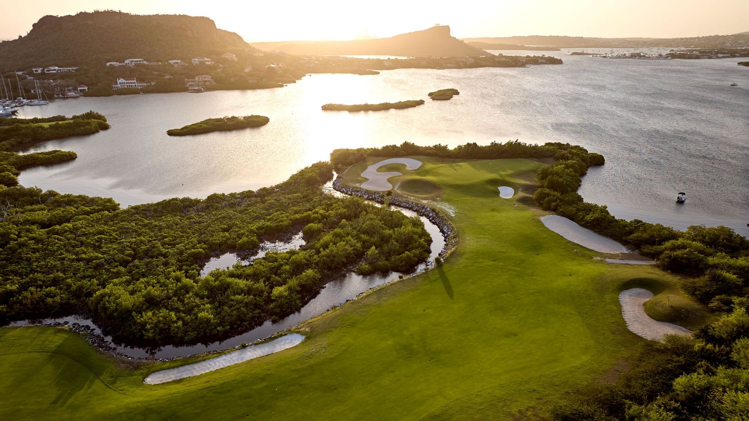 Panoramic aerial view of a coastal golf course with winding fairways surrounded by ocean water and green landscapes at sunset.