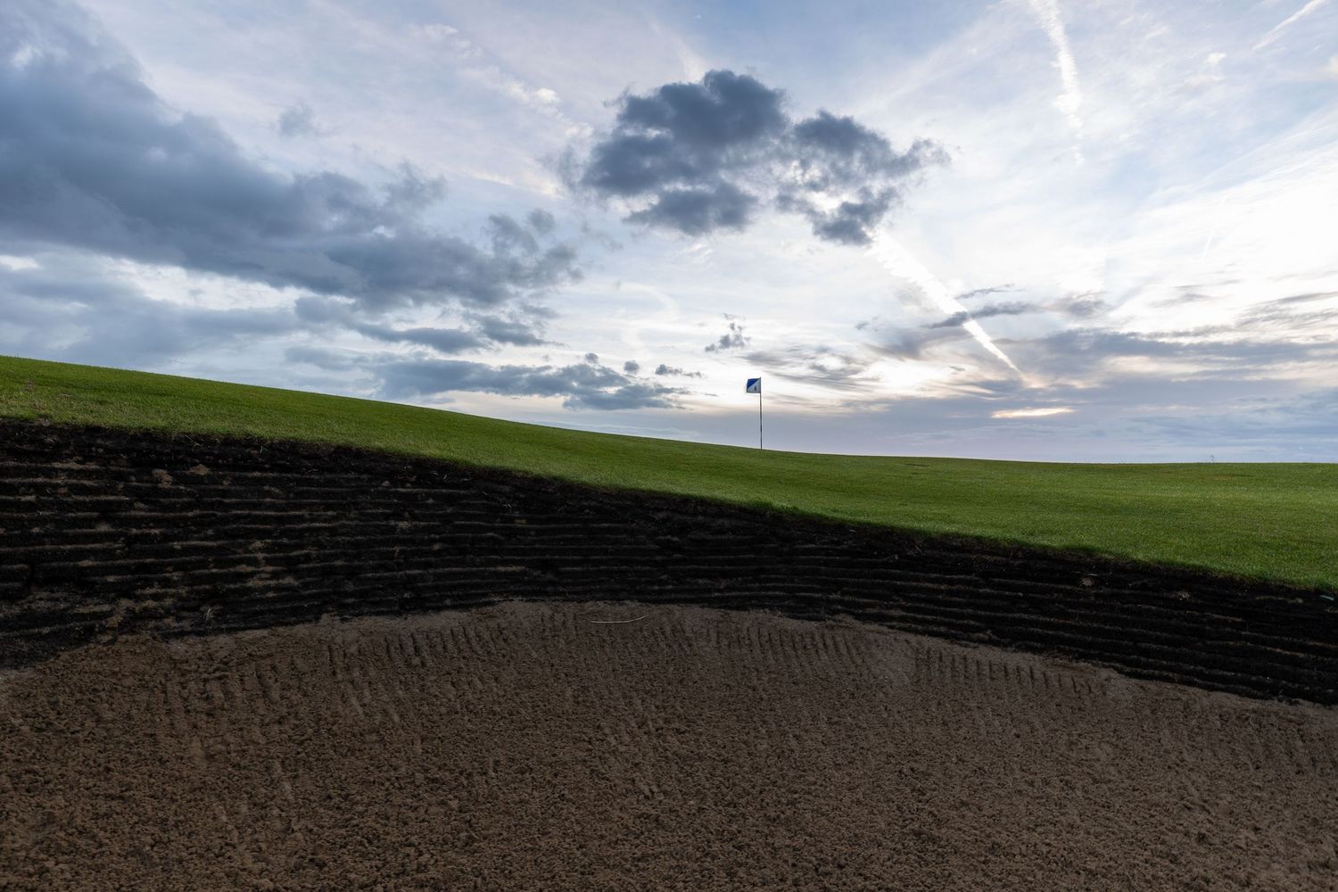 Golf course bunker with sand trap, green fairway, white flag pin, and dramatic cloudy sky at dusk.