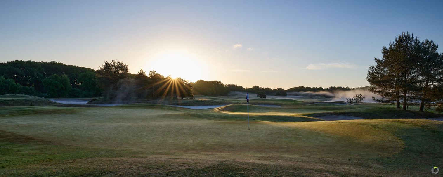 Panoramic view of a golf course fairway at sunset with golden light rays bursting through trees on the horizon.