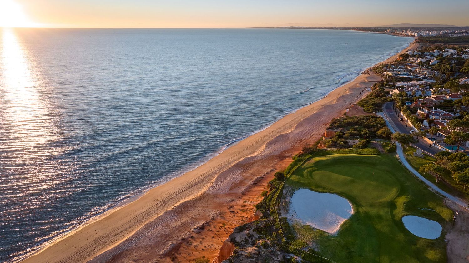 Aerial panoramic view of a coastal golf course with sandy beaches and blue ocean waters during golden sunset.