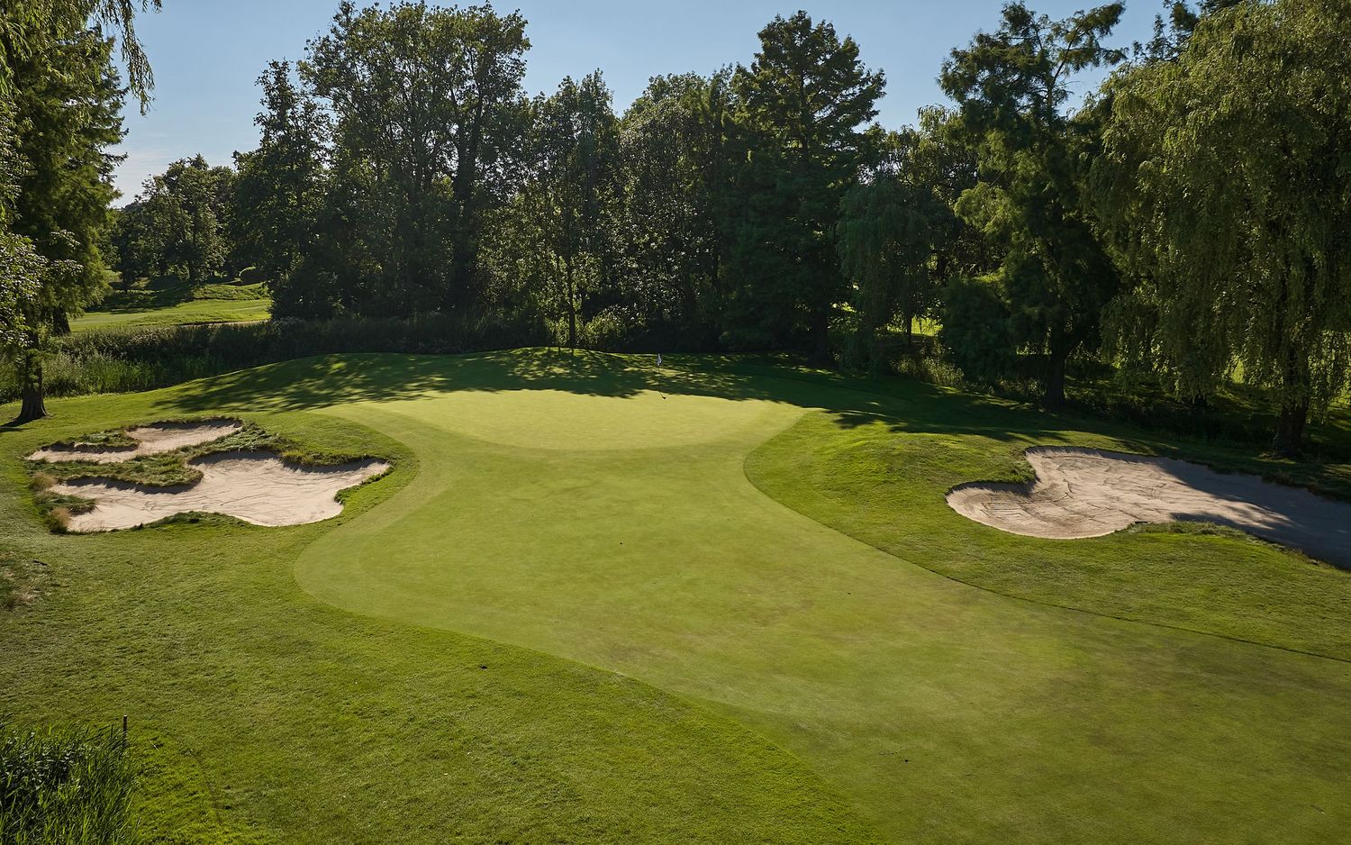 Golf course green with two sand bunkers on either side surrounded by tall trees on a sunny day.