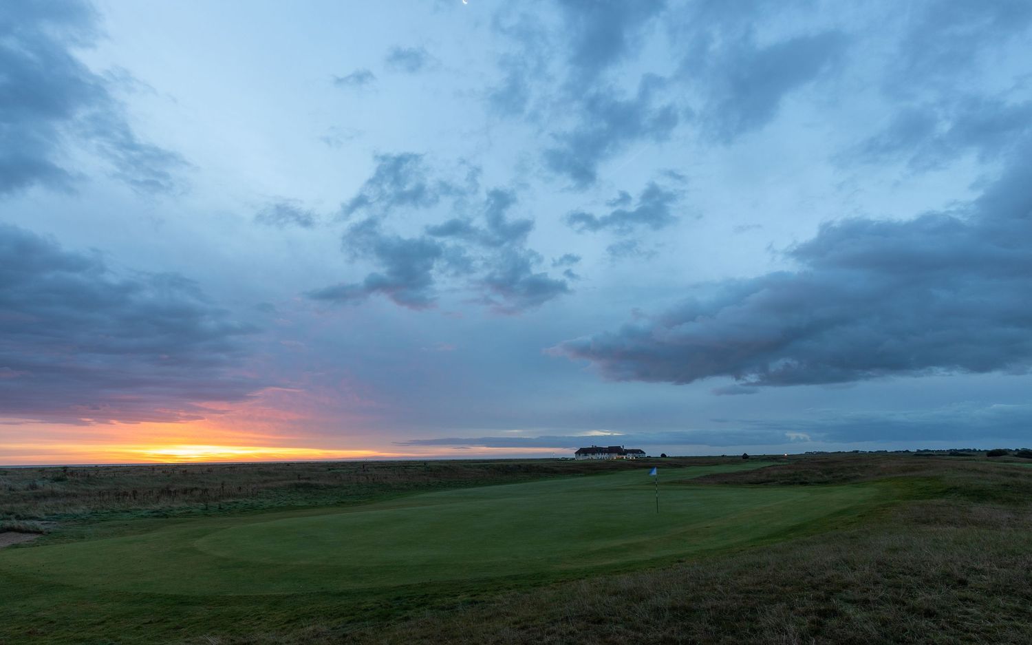 Dramatic sunset over a lush golf course with stormy clouds and warm horizon glow in an open landscape.