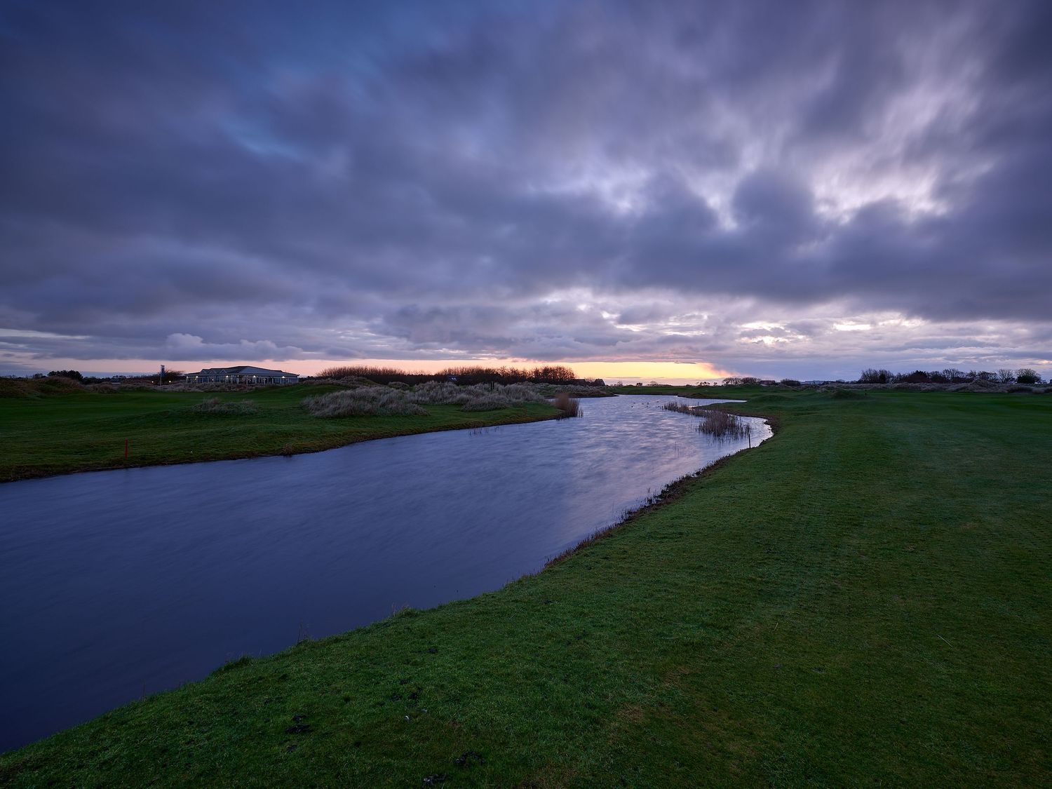 A dramatic sunset over a winding river with lush green banks under a moody cloudy sky in a rural landscape.